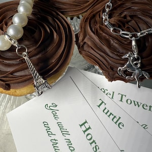 May include: An overhead shot features two cupcakes with chocolate frosting, and two silver charm bracelets. One bracelet has a pearl strand and an Eiffel Tower charm. The other has a horse charm. White cards with green text are in the foreground.