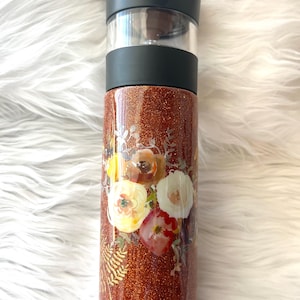 May include: A glittery copper-colored tumbler with a floral design. The tumbler has a black lid and a clear section. The floral design features watercolor-style flowers in shades of red, yellow, and white, with green foliage.