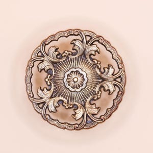 May include: An ornate, circular decorative plate with a detailed, symmetrical design. The plate features a central floral motif surrounded by radiating lines and stylized leaf patterns. The colour palette includes bronze, gold, and silver tones.