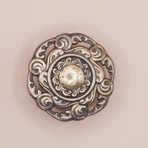 May include: An antique-style metal knob with an ornate, swirling design. The central part of the knob is raised and circular. The metal has a tarnished, aged appearance. Suitable for furniture restoration or decorative purposes.