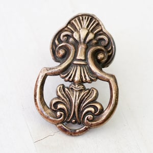 May include: Ornate brass cabinet pull with a decorative floral design. The pull has a ring shape and a detailed top.