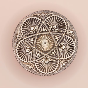 May include: A detailed, silver-coloured, round decorative object with an intricate, symmetrical design. The pattern features a central star shape surrounded by fan-like elements and ornate detailing. The object is set against a soft, pink background.