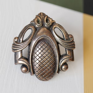 May include: A bronze-toned metal cabinet knob featuring an oval centre with a raised diamond grid pattern. The knob has an ornate, symmetrical frame with curved scrollwork details and small spherical accents at the base and sides.