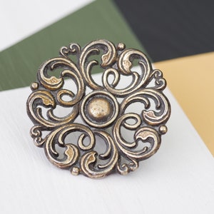 May include: A decorative, ornate, gold-toned metal knob with a circular design featuring intricate swirls and scrolls.