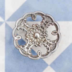 May include: Ornate silver drawer pull with a floral design. The decorative hardware features a central flower and swirling details. The pull is set against a blue and white checkered background.