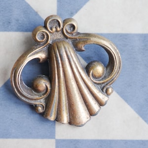 May include: An antique bronze-coloured drawer pull with a shell and scroll design. The hardware features a central fan-shaped element and curved, symmetrical arms. The aged finish adds a vintage aesthetic, suitable for furniture restoration or decorative projects.