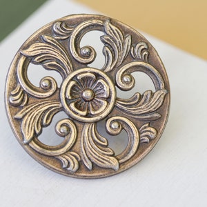 May include: A silver-toned metal button with a decorative floral design. The button has a raised centre with a flower shape and four swirling, leaf-like patterns around it.