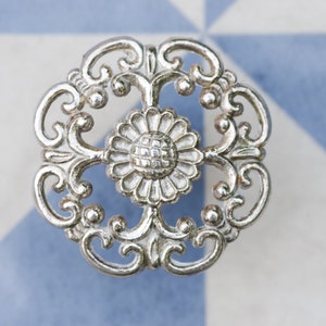 May include: A silver decorative knob with a floral design. The knob features a central flower with a textured centre and is surrounded by intricate scrollwork.