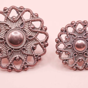 May include: Two ornate copper-coloured metal buttons with a circular design and intricate details. The larger button has a diameter of approximately 5 cm, while the smaller button is about 2.5 cm in diameter.