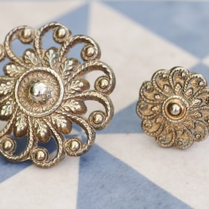May include: Two ornate silver-toned metal drawer pulls with intricate floral designs. The larger pull has a circular centre with a raised, textured surface. The smaller pull has a similar design but is smaller in size.