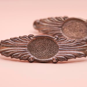 May include: Two antique silver-toned drawer pulls with an ornate design. Each pull features an oval centre with a textured surface, framed by a detailed, flowing pattern. The background is a soft pink.