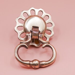 May include: Vintage brass-effect drawer pull featuring a floral backplate and a curved handle. The hardware is set against a pale pink background, highlighting its antique design.