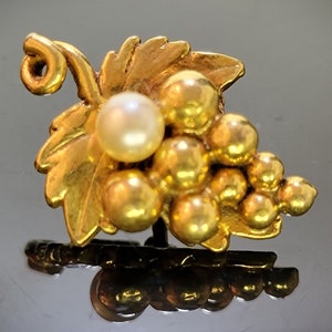May include: A gold-colored brooch in the shape of a grape cluster with leaves. The brooch features a pearl and several gold-colored spheres, and is approximately 1 inch in length.