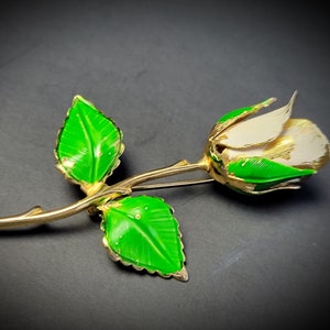Puede incluir: Un broche de rosa de tono dorado con pétalos de esmalte blanco y hojas de esmalte verde.