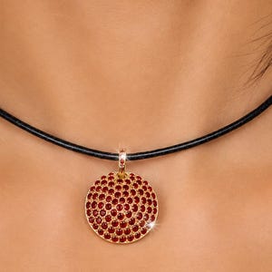 Puede incluir: Un collar de cordón negro con un colgante circular en un engaste de tono oro rosa. El colgante está densamente incrustado con numerosas piedras preciosas redondas de color rojo oscuro que crean una superficie texturizada y brillante.