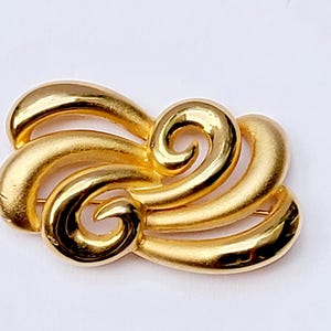 Puede incluir: Broche dorada con un diseño abstracto y ondulado. La broche presenta elementos lisos y curvos que crean una sensación de movimiento. Este accesorio clásico añade un toque de elegancia a cualquier atuendo.
