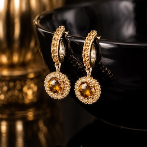 May include: A pair of gold hoop earrings featuring a dangling circular pendant. Each pendant contains a central faceted amber-colored gemstone surrounded by a halo of small clear stones. The hoop portion is also encrusted with small clear stones.