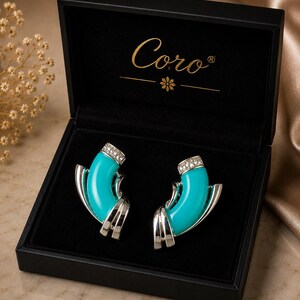 May include: A pair of silver-tone earrings featuring curved, bright turquoise enamel inserts and clear rhinestone accents at the top. The earrings are presented inside a black hinged jewelry box with the gold text Coro printed on the lid.