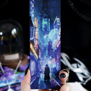 Peut inclure: Un marque-page représentant une ville futuriste avec des enseignes au néon et une silhouette solitaire en manteau sombre. La palette de couleurs est dominée par le bleu et l'orange, créant une esthétique cyberpunk. Le marque-page est tenu dans une main.