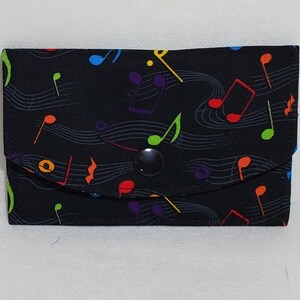 May include: A black fabric wallet with a flap closure and a black snap. The wallet is decorated with colorful musical notes and staff lines. The notes are in shades of red, yellow, green, blue, and purple.