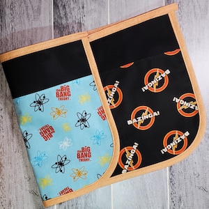 May include: A black and blue fabric apron with a pocket on the front. The apron has a pattern of the Big Bang Theory logo and the word "Bazinga!" repeated on the fabric.