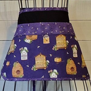 May include: Purple and black apron with a bee and honeycomb pattern. The apron has a black waistband and a purple and white honeycomb pattern. The apron has the text "Home Sweet Home" on it.