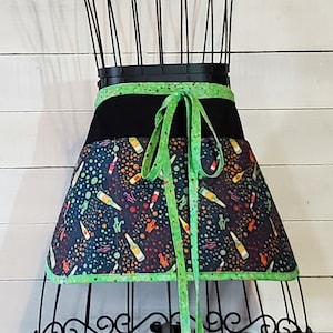 May include: A black and green apron with a colorful print of bottles and polka dots. The apron has a green tie and a black waistband.