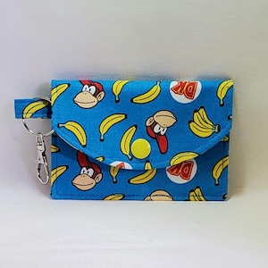 May include: A blue wallet with a flap closure, featuring a pattern of cartoon monkeys, bananas, and DK logos. The wallet has a wrist strap with a silver-colored key ring attached.