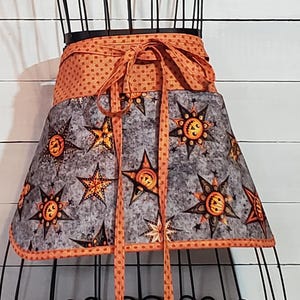 May include: A Halloween-themed apron with a gray and orange color scheme. The apron features a gray patterned fabric with star and sun designs, and an orange polka dot waistband and ties. The apron is displayed on a black mannequin.