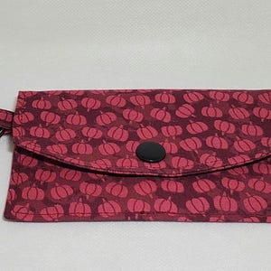 May include: A burgundy fabric wallet with a repeating pattern of pink pumpkins. The wallet has a flap closure with a black button and a silver key ring with a clasp. The wallet is designed to hold cards and keys.