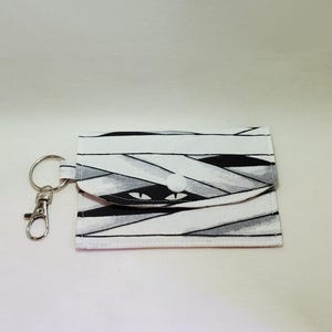 May include: A white, black, and gray mummy-themed keychain wallet with a silver key ring and clasp. The wallet features a button closure and a mummy face design.