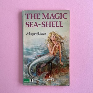 May include: A vintage paperback book titled "THE MAGIC SEA-SHELL" by Margaret J. Baker. The cover features an illustration of a mermaid with long blonde hair, sitting on a rock near the ocean. The book has a pink background.