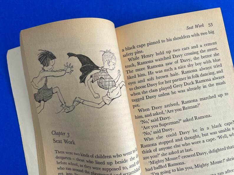 以下が含まれることがあります： イラストとテキストが掲載された開いた本。左ページには、2人の子供が走っている白黒の絵が描かれています。右ページには「Seat Work」というタイトルと番号53のテキストが含まれています。