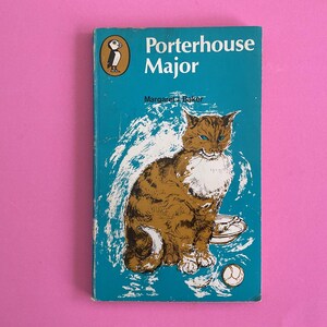 May include: Vintage book cover titled "Porterhouse Major" by Margaret J. Baker. The cover is teal with a brown and white illustration of a cat. The Penguin logo is in the upper left corner. A white and blue background surrounds the cat.