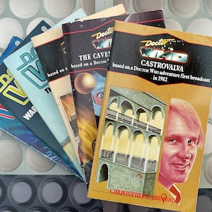 May include: A collection of five Doctor Who paperback books, all featuring the Doctor Who logo and the title of the book. The books are arranged in a fan shape, with the top book being "Castrovalva" and the bottom book being "The Caves of Androzani".