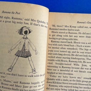 以下が含まれることがあります： テキストと、口を開けた女の子の白黒イラストが描かれた開いた本。「Ramona the Pest」と「Ramona's Great Day」と書かれています。