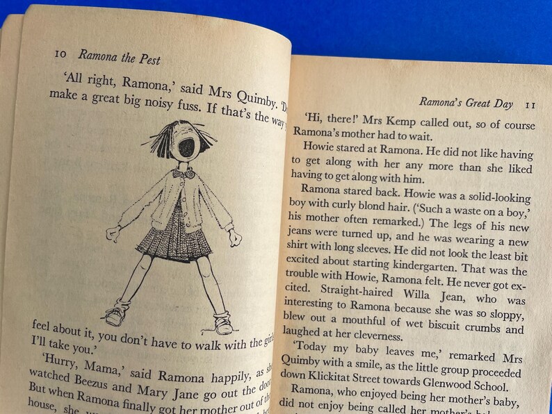以下が含まれることがあります： テキストと、口を開けた女の子の白黒イラストが描かれた開いた本。「Ramona the Pest」と「Ramona's Great Day」と書かれています。