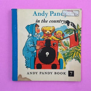 May include: Vintage children's book cover titled "Andy Pandy in the Country." The cover features illustrations of Andy Pandy, a teddy bear, and a girl on a red and black train. A bird perches on a flag. The book is number 7.