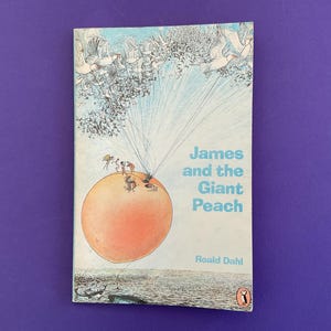May include: A paperback book titled "James and the Giant Peach" by Roald Dahl. The cover features an illustration of a large peach with figures on top, suspended in the air by strings attached to birds. The background is a light blue sky.