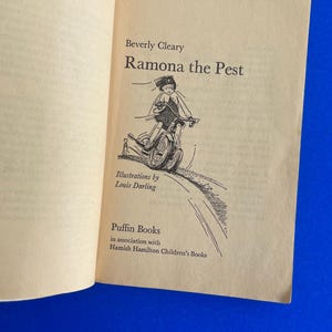 以下が含まれることがあります： ベバリー・クリアリー著「Ramona the Pest」のタイトルが書かれた開いた本のページ。ルイ・ダーリングによるイラストは、自転車に乗る子供を描いています。パフィンブックスとハミッシュ・ハミルトン・チルドレンズ・ブックスの共同出版。