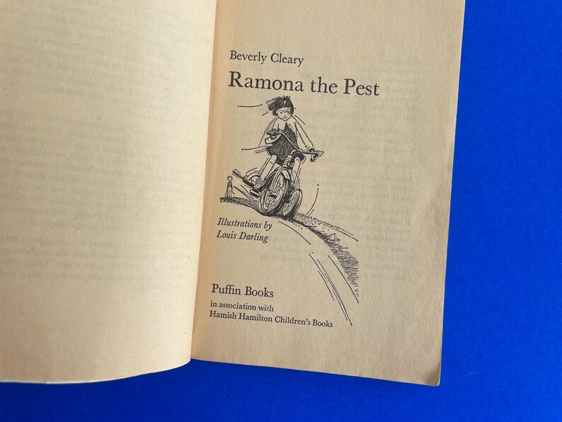 以下が含まれることがあります： ベバリー・クリアリー著「Ramona the Pest」のタイトルが書かれた開いた本のページ。ルイ・ダーリングによるイラストは、自転車に乗る子供を描いています。パフィンブックスとハミッシュ・ハミルトン・チルドレンズ・ブックスの共同出版。