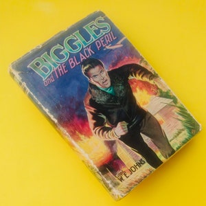 May include: A vintage paperback book titled "Biggles and The Black Peril" by Capt. W.E. Johns. The cover features a man in a leather jacket running through a fiery scene.