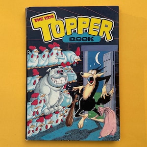 May include: The 1974 Topper Book is a hardback book featuring a colourful comic illustration on the front cover. The artwork depicts a large white bulldog surrounded by many white chickens, while a black wolf holding a club stands in a doorway.