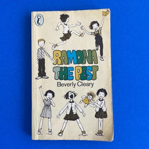 以下が含まれることがあります： ベバリー・クリアリー著「Ramona the Pest」というタイトルのヴィンテージペーパーバック。表紙には子供たちのイラストとカラフルな文字でタイトルが描かれています。左上にはペンギンのロゴがあります。