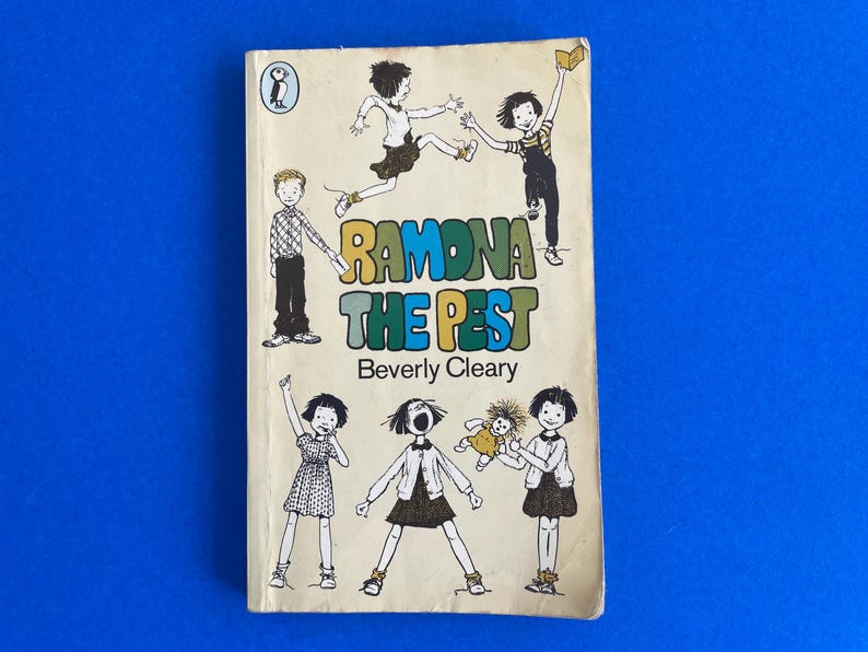 以下が含まれることがあります： ベバリー・クリアリー著「Ramona the Pest」というタイトルのヴィンテージペーパーバック。表紙には子供たちのイラストとカラフルな文字でタイトルが描かれています。左上にはペンギンのロゴがあります。