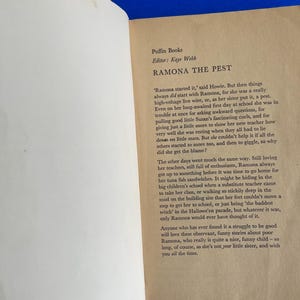 以下が含まれることがあります： Puffin Booksの「Ramona the Pest」からのテキストが書かれた開いた本。ページはオフホワイトで、黒い文字が書かれています。本は青い表面に開かれています。