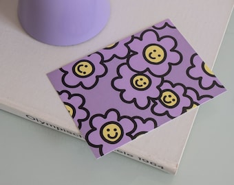 Könnte beinhalten: Eine lilafarbene Lampe mit einem breiten, flachen Schirm steht auf einer weißen Oberfläche. Eine lilafarbene Karte mit einem sich wiederholenden Muster aus schwarz umrandeten gelben Smiley-Gesichtern in lilafarbenen Blumen liegt auf der Oberfläche.