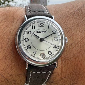 May include: A silver-toned Raketa wristwatch with a light-colored face and black hands. The watch has a brown leather strap with white stitching. The watch face displays the numbers 1 through 12, and the date. The watch is on a wrist.