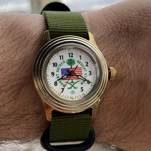May include: A gold-tone wristwatch with a white face and black hands. The watch face features a green and white logo with the text "Desert Storm" and "Desert Shield". The watch has a green nylon strap.