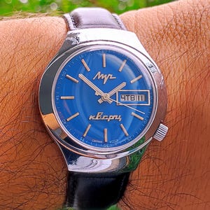 May include: A silver-tone wristwatch with a vibrant blue dial, featuring silver hour markers, silver hands, and a date window displaying Cyrillic text. The watch has a black leather strap and a polished metal case with a side crown.
